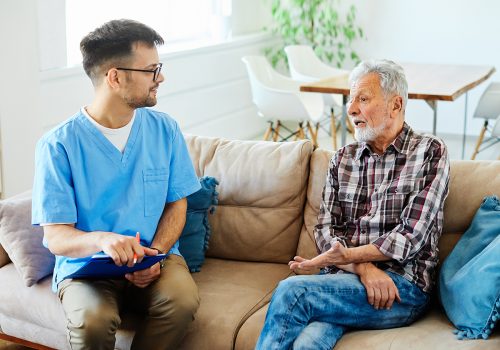 Doctor or nurse caregiver talking with senior man on sofa at home or nursing home